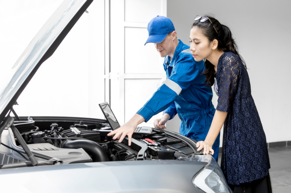 A mechanic shows a lady who is his client the inside of her car's engine while reading car data on his laptop.