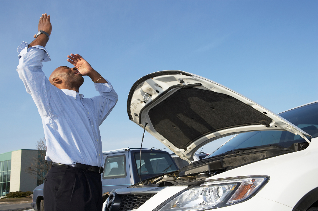 An angry, frustrated man throws his arms up in the air in frustration as his white car is broken down with its bonnet raised.